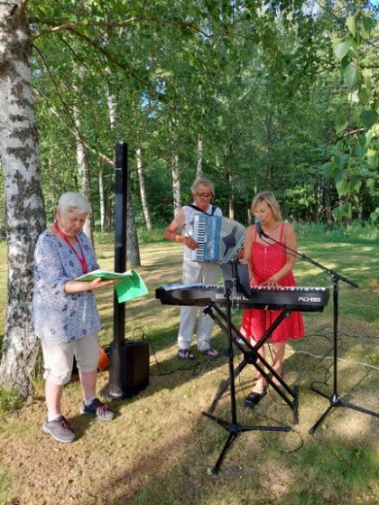 Tre personer spelar musik utomhus bland träd, en med keyboard, en med dragspel och en med papper.