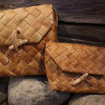 Två handgjorda väskor av flätad näver ligger på stenar framför en trävägg.