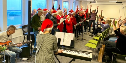 En kör med tomteluvor sjunger och spelar instrument inför en publik i ett ljust rum.