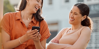 Två unga kvinnor står utomhus och skrattar tillsammans, en håller en mobiltelefon.