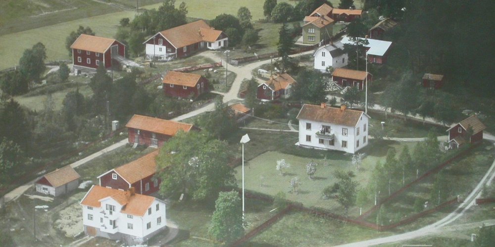 Flygfoto över en liten by med röda och vita hus, omgiven av grönska och åkrar.