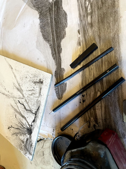 Blyertspennor, kol och ett bläckburk ligger på ett bord med teckningar av landskap i svartvitt.