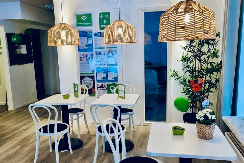 Ljust och modernt café med vita stolar, träbord, växter och flätade lampor i taket.