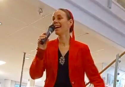 Person i röd kavaj håller mikrofon och talar inomhus, med modern inredning i bakgrunden.