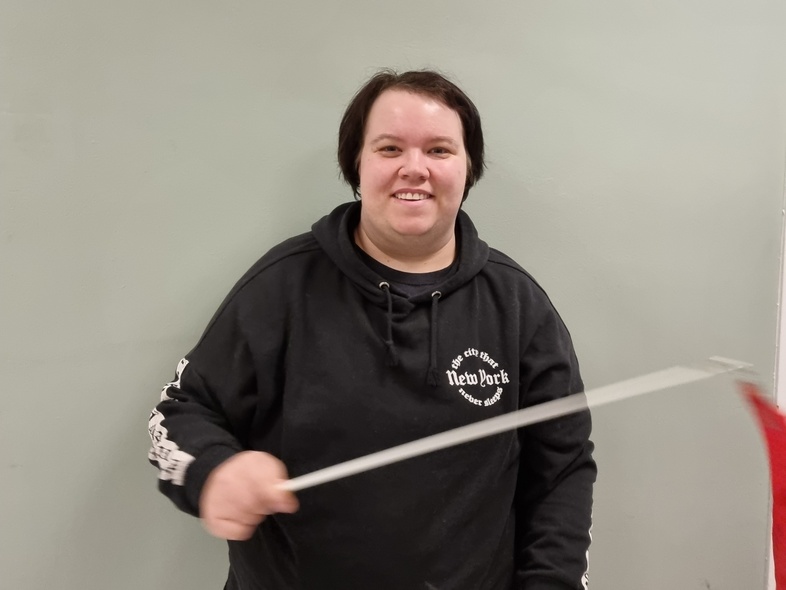 Person i svart hoodie med texten 'New York' håller ett rött band på pinne mot ljusgrå vägg.