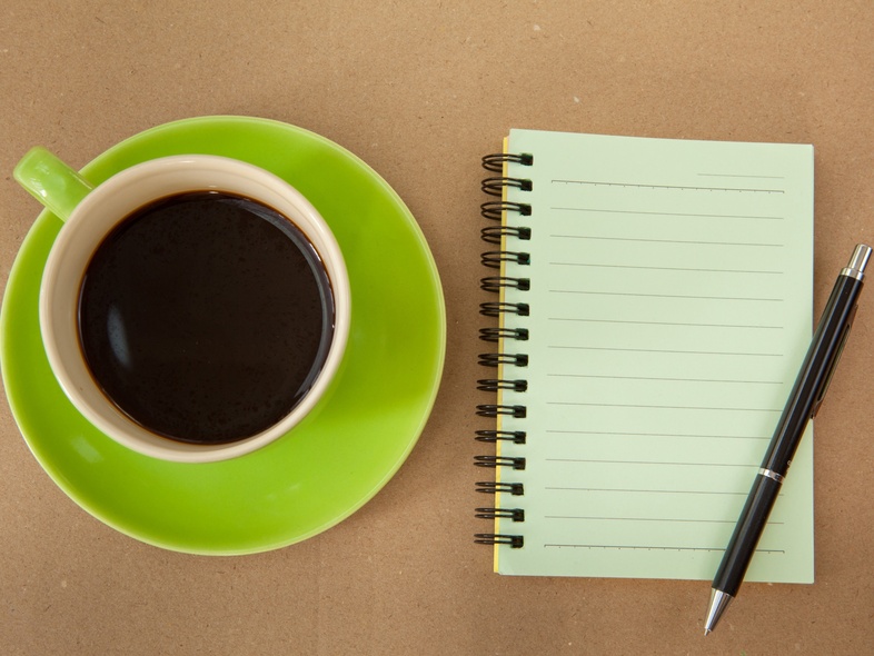 En kopp svart kaffe på ett grönt fat bredvid ett tomt spiralblock och en penna på ett brunt bord.