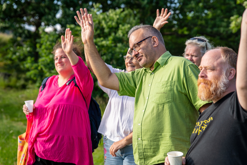 En grupp vuxna personer står utomhus och räcker upp händerna, några håller i muggar. Soligt väder och grönska i bakgrunden.