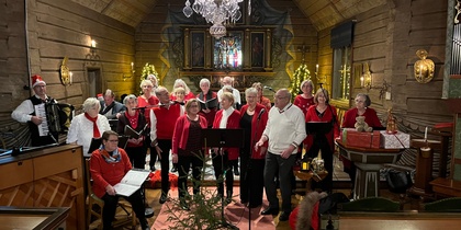 En kör sjunger i en träkyrka dekorerad för jul, med musiker och publik närvarande.