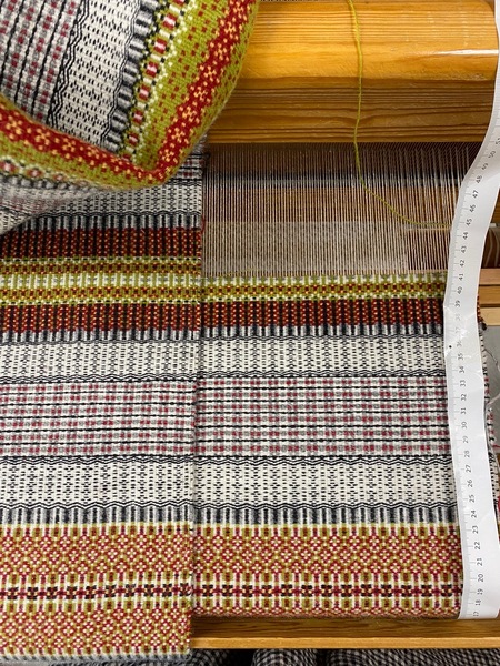 Närbild av en vävstol med färgglad väv i rött, gult, svart och vitt samt ett måttband vid sidan.
