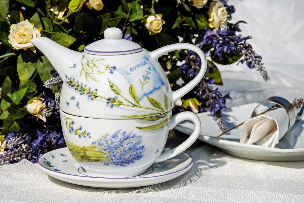 En tekanna och kopp med lavendelmotiv står på ett fat, omgiven av blommor och en servett på tallrik.