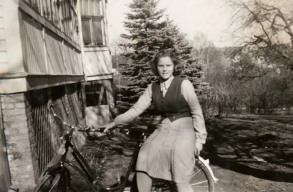 Kvinna sitter på en cykel utomhus bredvid ett hus, med träd i bakgrunden, i en svartvit bild.