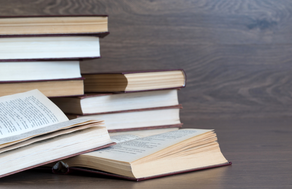 Flera böcker ligger och staplas på ett träbord, några är öppna och andra stängda.