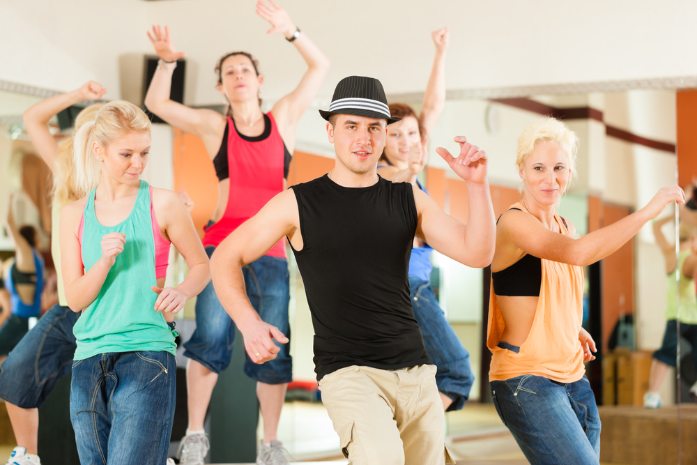 En grupp människor dansar energiskt i ett ljust träningsrum, ledda av en man med hatt.