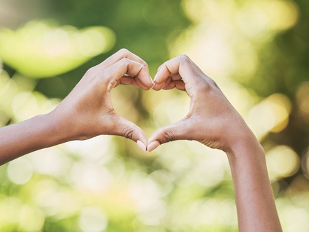 Två händer formar ett hjärta mot en suddig, grön bakgrund utomhus.