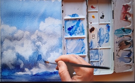 En hand målar moln på ett papper med akvarellfärger och pensel, färgpalett syns bredvid.