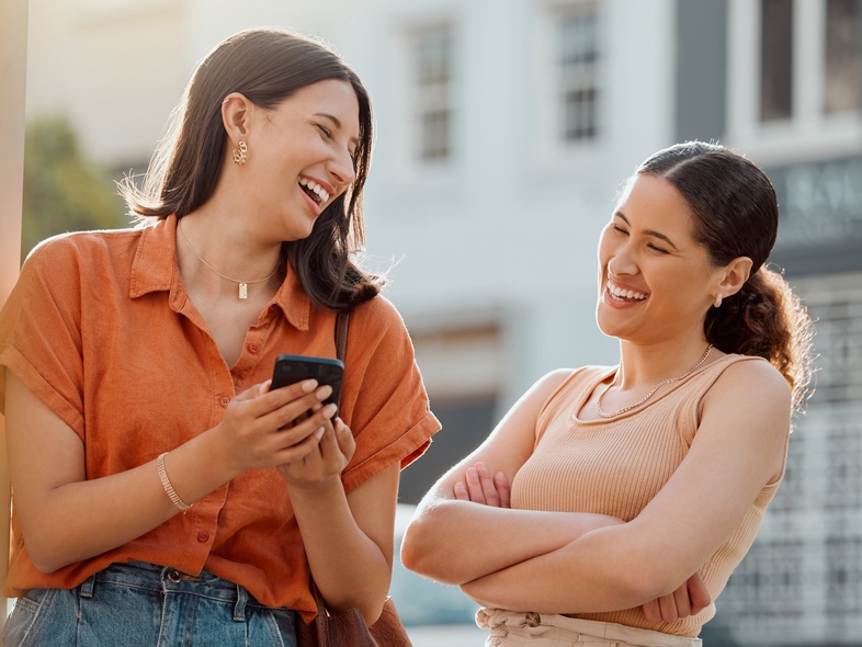 Två unga kvinnor står utomhus och skrattar tillsammans, en håller en mobiltelefon.
