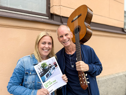 Två leende personer står utomhus, en håller en gitarr och den andra ett papper med bilder på ett kök.
