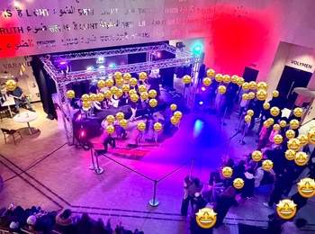 Vy ovanifrån av ett evenemang inomhus med många människor, scen, färgglad belysning och text på väggen.