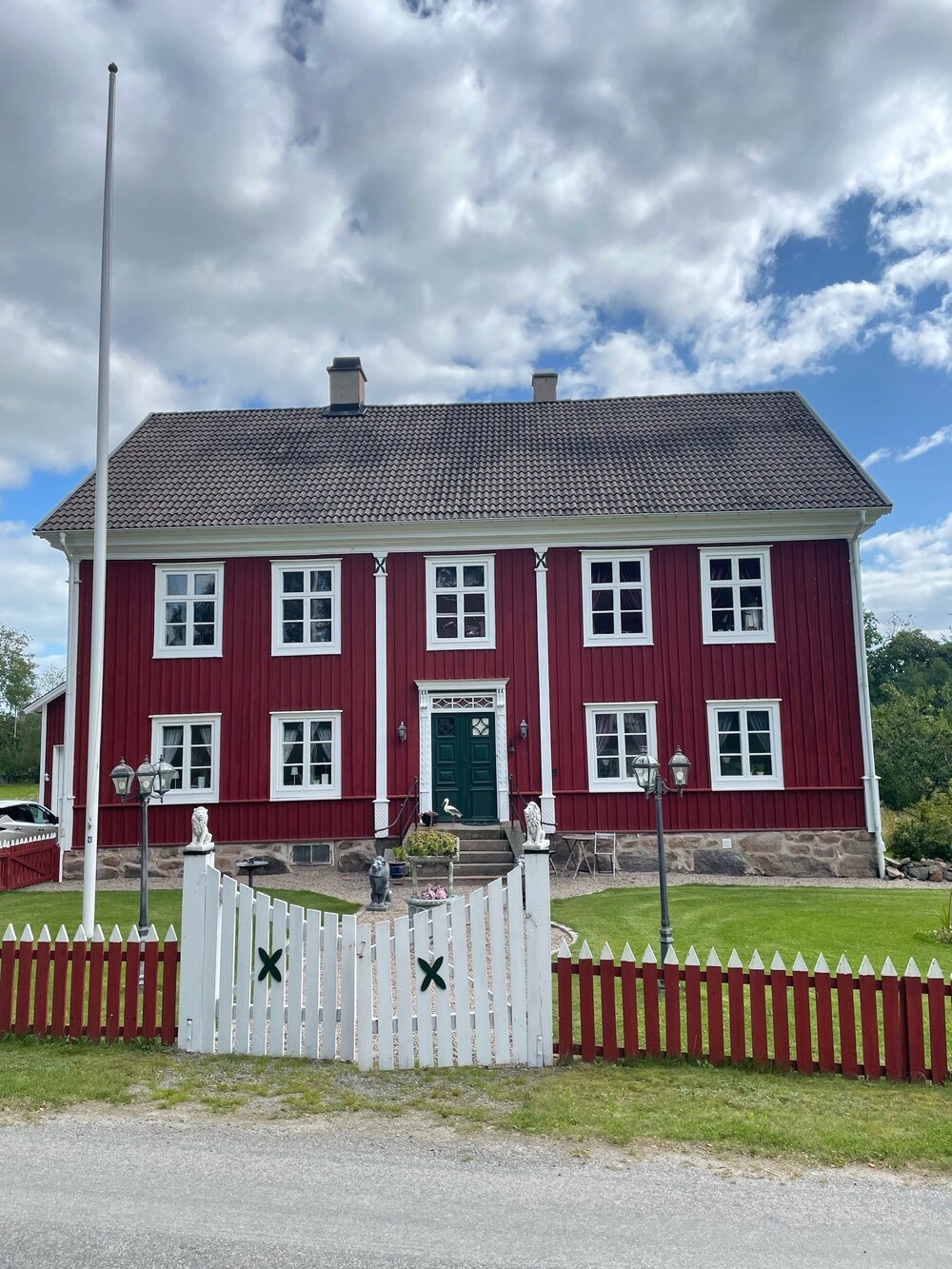Röd trävilla med vita knutar, staket och grind framför, under en delvis molnig himmel.