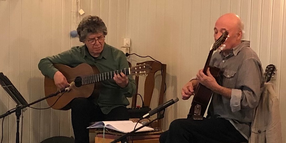 Två personer spelar akustisk gitarr på en liten scen i ett mysigt rum med vita träväggar.