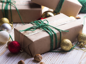 Inslagna paket med brunt papper och grönt snöre, omgivna av julkulor och nötter på ett trägolv.