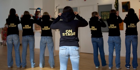Sju personer står med ryggen mot kameran, alla bär svarta huvtröjor med texten 'ON STAGE' och blå jeans i ett ljust rum.