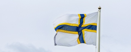 Flagga med blått kors och gul kant på vit bakgrund vajar mot en molnig himmel.