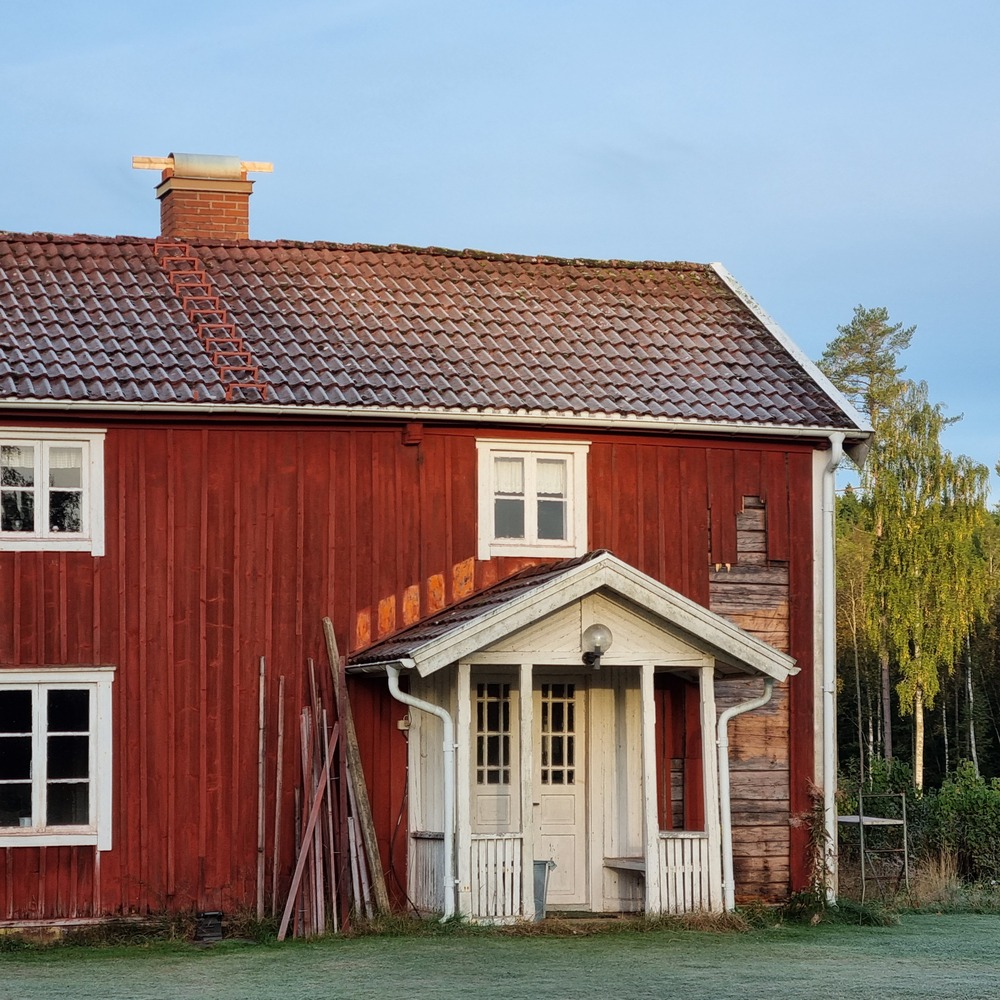 Röd trähus med vita detaljer och tegeltak, omgiven av grönska och morgondagg på marken.