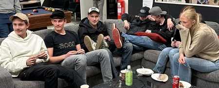 En grupp unga vuxna sitter i en soffa runt ett svart soffbord med drycker och tallrikar i ett vardagsrumsliknande rum.