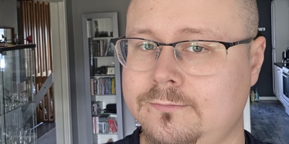 Man med glasögon och kort skägg tar en selfie i ett modernt vardagsrum med grå väggar.