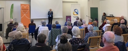 En person står och talar inför en sittande publik i en ljus sal med träbjälkar och stora lampor i taket.