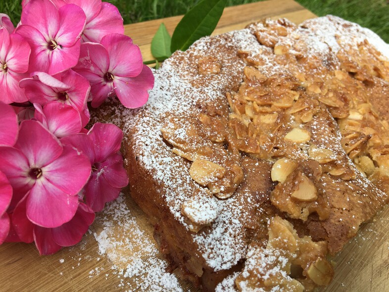 En mandelkaka med florsocker på ett träfat, dekorerad med rosa blommor bredvid.