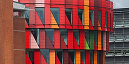 Färgglad, modern byggnad med röd, orange och gul fasad i geometriska mönster, omgiven av träd och andra byggnader.