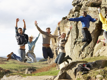 Sex personer hoppar glada i luften vid en klippa i ett naturskönt landskap.
