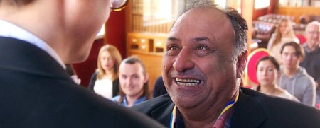 En man med medalj ler stort mot en person i kostym, med en publik i bakgrunden i en ljus sal.