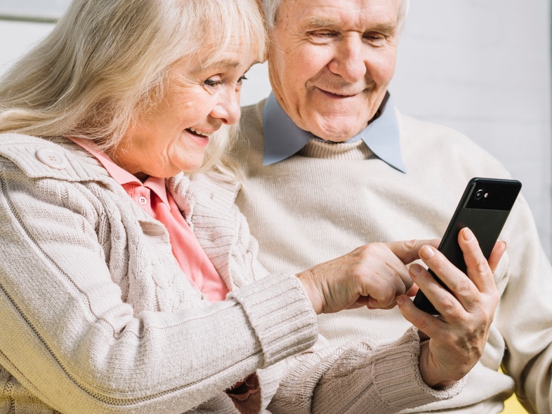 Ett äldre par sitter tillsammans i en soffa och tittar leende på en smartphone.