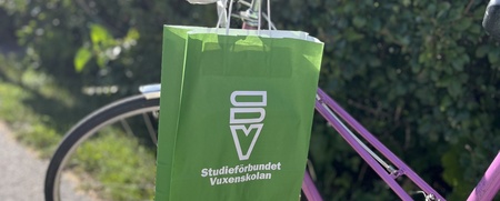 En grön papperspåse med Studieförbundet Vuxenskolans logotyp på hänger på styret till en rosalila cykel som står på en solig cykelväg bredvid en häck.