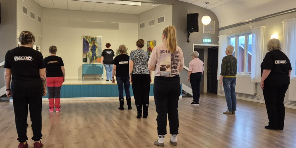 En grupp personer står i ett dansrum och följer en instruktör på en liten scen längst fram.