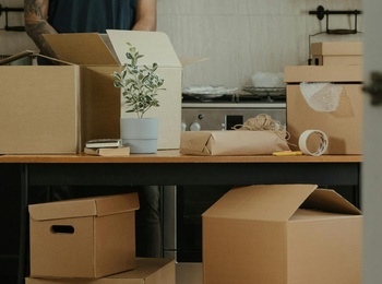 En person packar upp flyttkartonger i ett kök, sedd genom en öppen dörr.