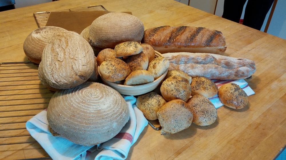 Olika sorters hembakat bröd och bullar ligger på ett träbord, några på en handduk och andra i en korg.