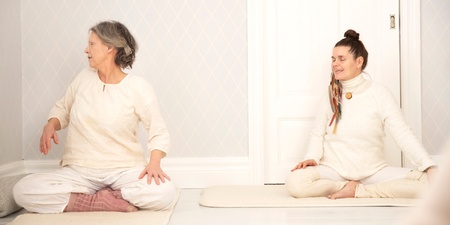 Två personer sitter på yogamattor i ett ljust rum och gör en sittande vridning.