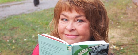 Kvinna med ljust hår sitter utomhus och ler medan hon håller upp en bok med titeln 'Sköna Maj'.