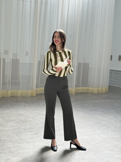 Person i randig tröja och svarta byxor står i ett ljust rum med gardiner och håller ett papper.