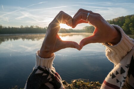 Två händer formar ett hjärta mot en sjö i soluppgång, med skog i bakgrunden.