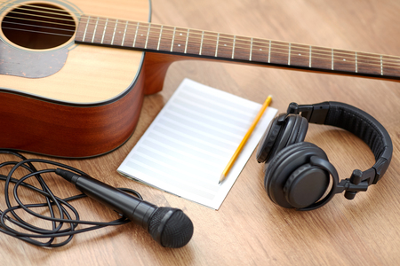 Akustisk gitarr, mikrofon, hörlurar, notpapper och penna på ett trägolv, redo för låtskrivning.