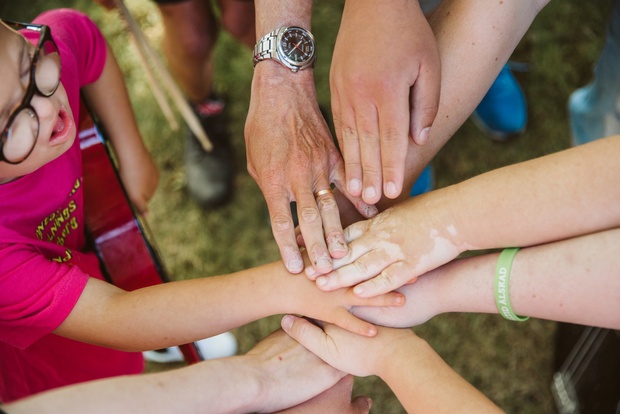 Flera händer i olika hudtoner läggs ovanpå varandra i en gemensam gest av samarbete utomhus.