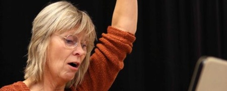 Kvinna med ljust hår och glasögon sitter vid ett piano och räcker upp ena armen, svart bakgrund.