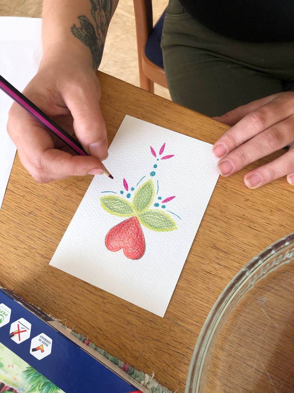 En person ritar ett färgglatt blommönster på ett vitt papper med färgpennor vid ett träbord.