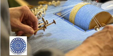 Närbild av händer som arbetar med knyppling på en blå knyppeldyna, med flera trådar och pinnar.