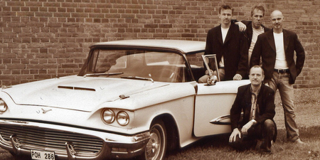 Fyra män i svarta kavajer står bredvid en äldre vit bil framför en tegelvägg